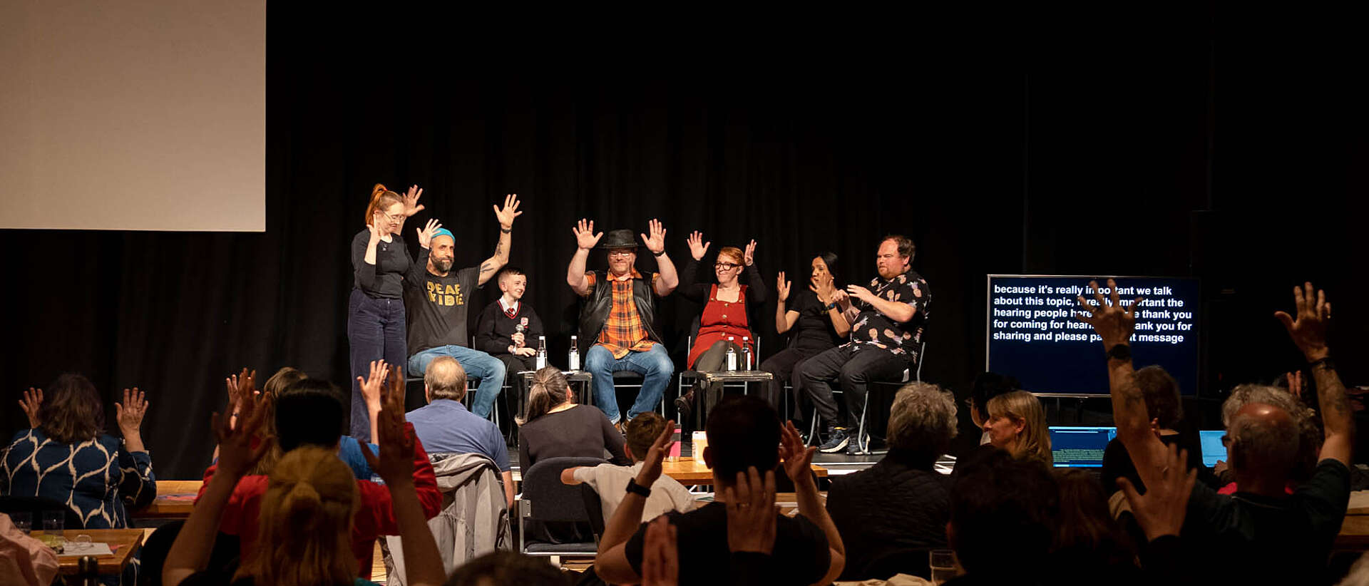 A group of people on stage applaud via BSL, the crowd watching them also applaud via BSL and through clapping.