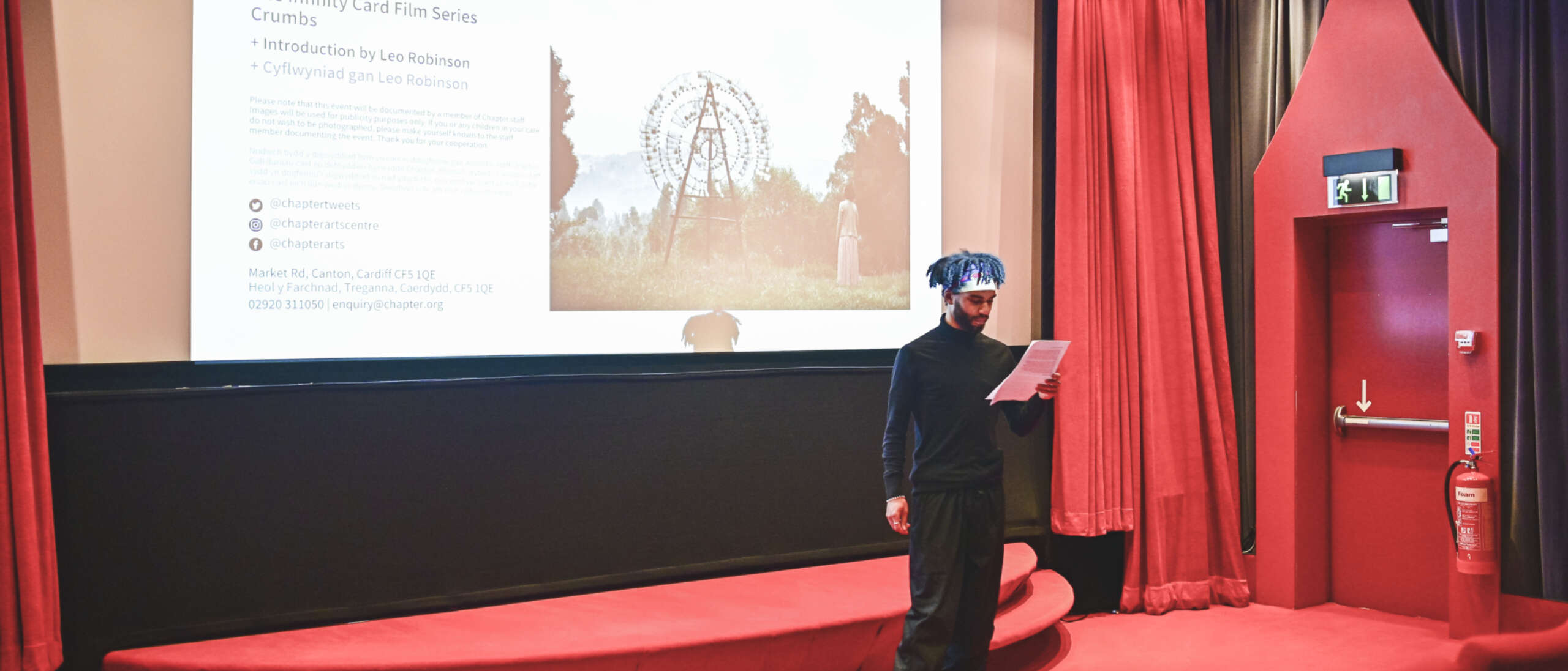 Artist Leo Robinson stands in front of a small cinema with red seats and doorways and reads off a piece of paper to the audience.