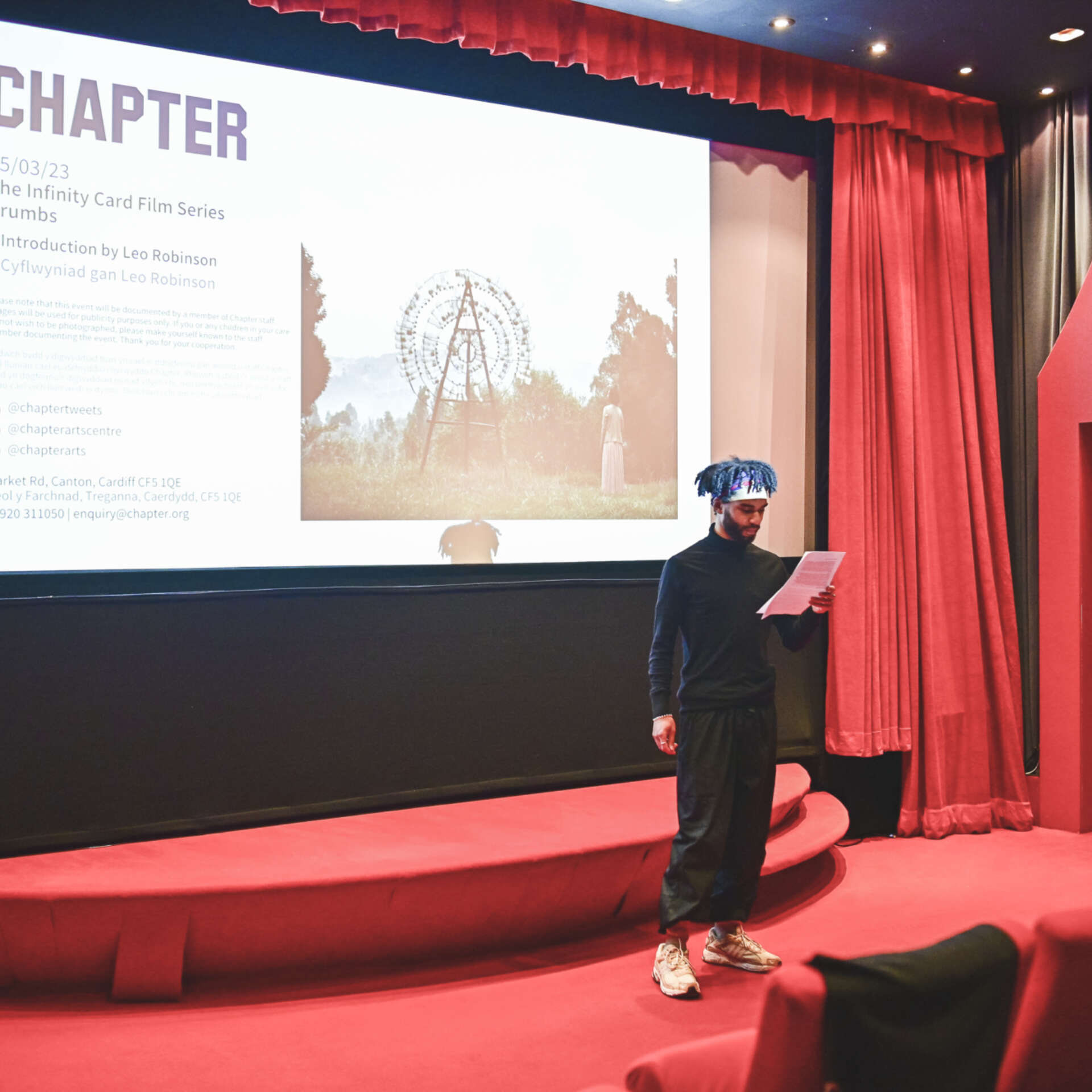 Artist Leo Robinson stands in front of a small cinema with red seats and doorways and reads off a piece of paper to the audience.