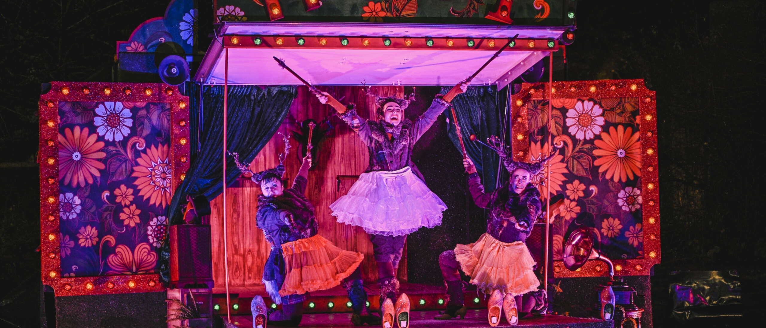 Theatr Iolo and Kitsch & Sync Colelctive perform on a stage at Chapter. In a marquee style with large floral boxes to the side, the performners are extending their arms in a row of three, the middle performer with their arts outstreched above everyone else.