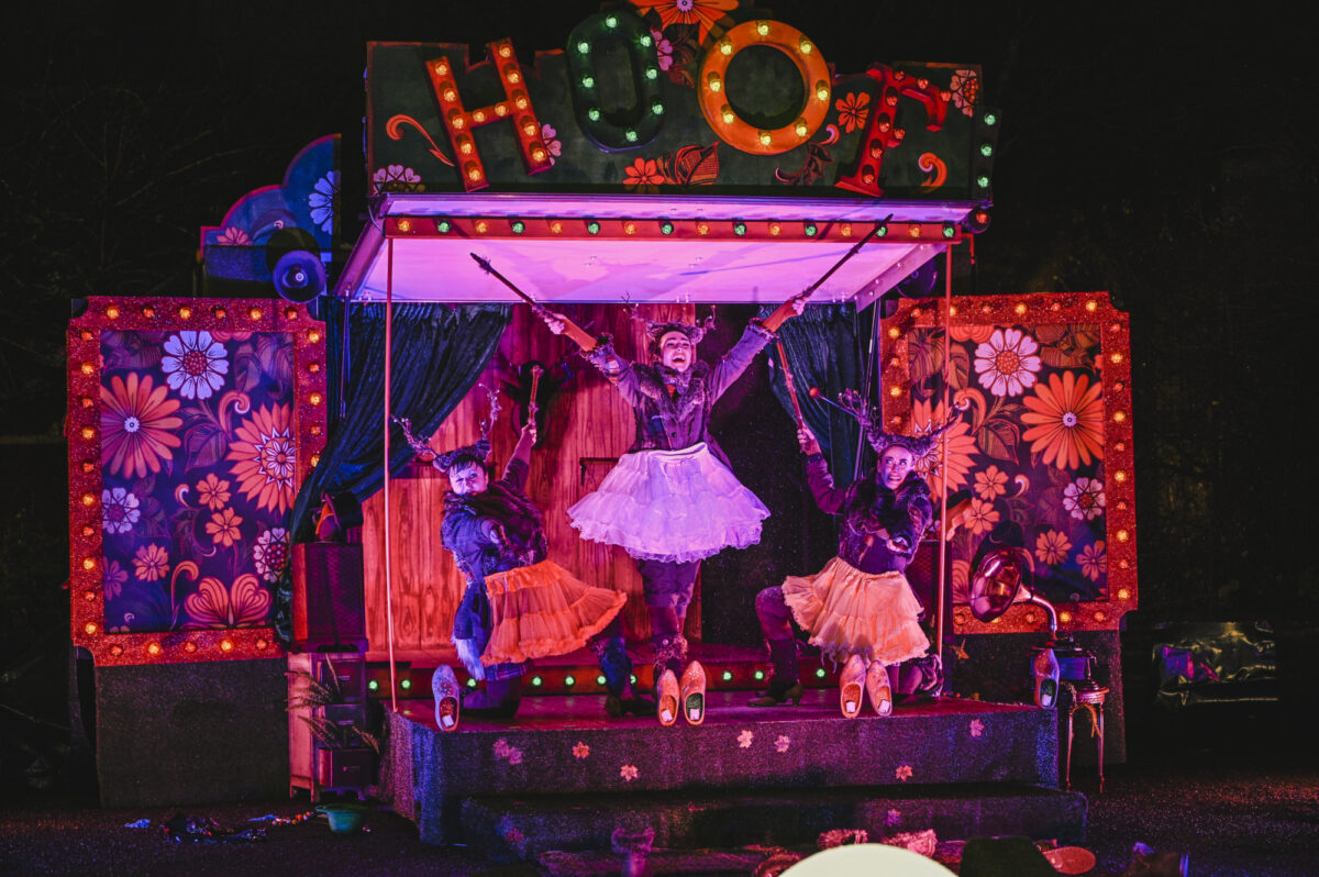 Theatr Iolo and Kitsch & Sync Colelctive perform on a stage at Chapter. In a marquee style with large floral boxes to the side, the performners are extending their arms in a row of three, the middle performer with their arts outstreched above everyone else.
