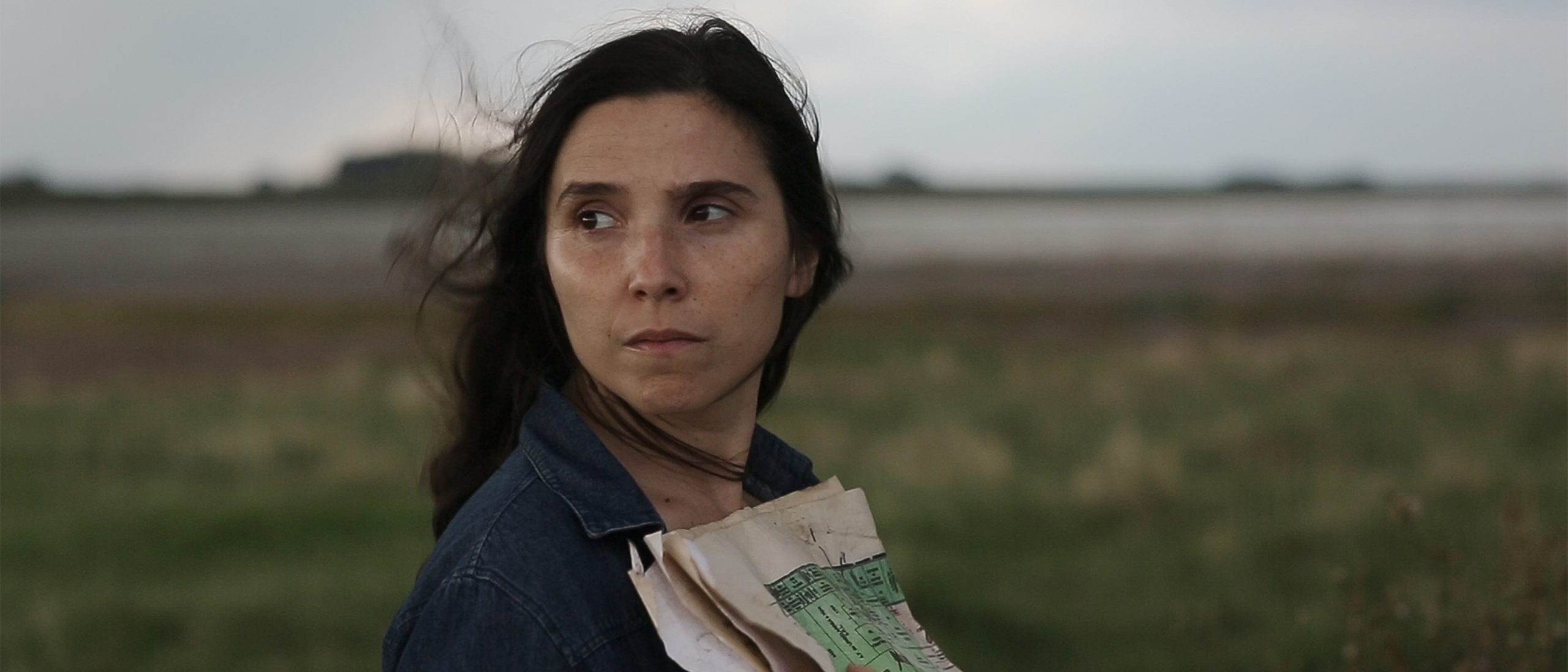 Woman with long dark brown straight hair looks over her right shoulder. She stands in the middle of a green field with the sea in the background and holds a pile of used papers to her chest. Still image from the film Trenque Lauquen I & II.